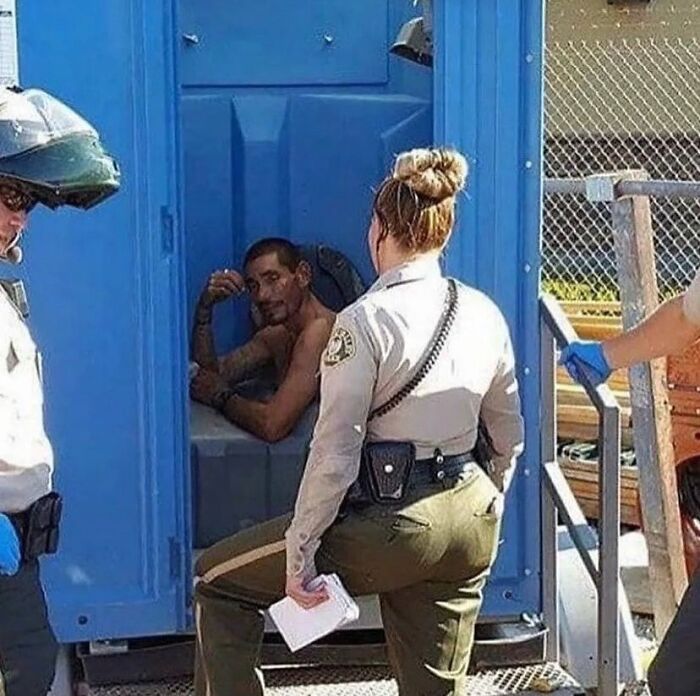 Hombre dentro de baño portátil rodeado por oficiales uniformados en imagen maldita difícil de explicar.