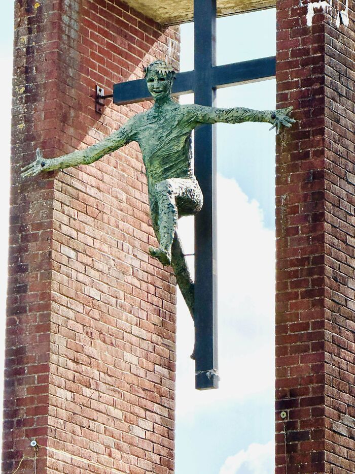 Escultura aterradora e inquietante de figura humana verde colgada frente a cruz negra entre paredes de ladrillo rojo.