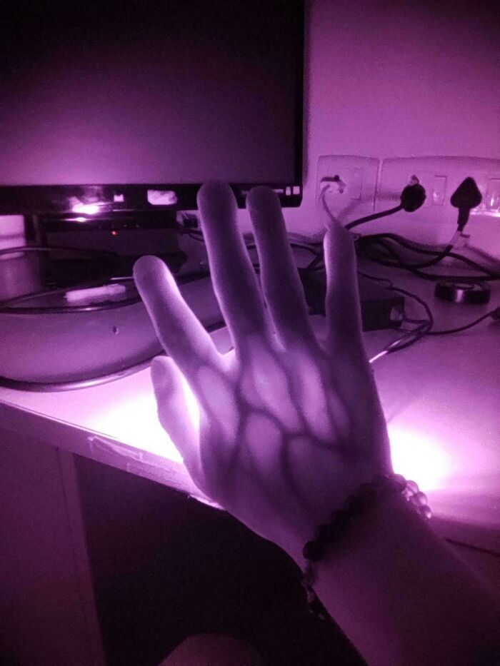 Mano iluminada con luz morada creando sombras interesantes en un escritorio con enchufes y cables al fondo.