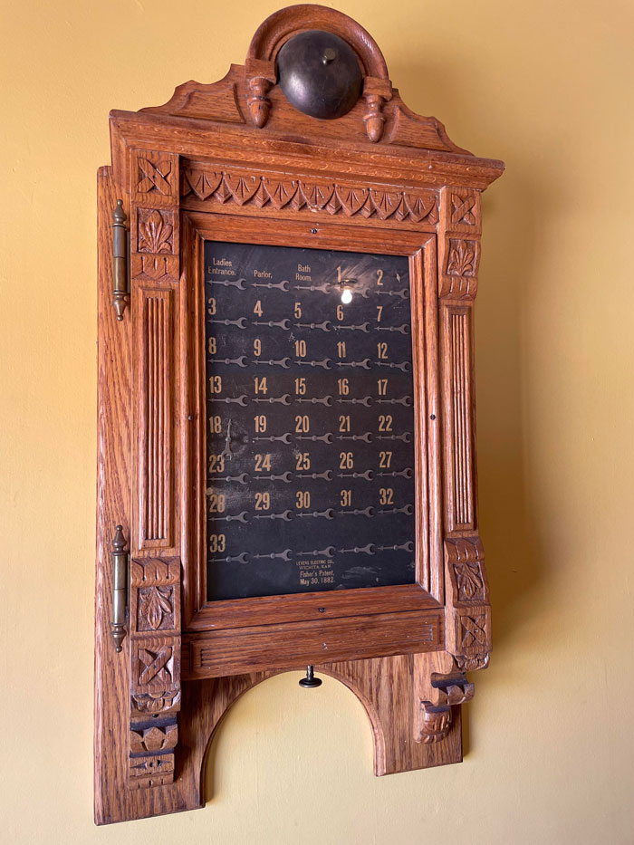 Máquina de madera colgada en la pared de un edificio histórico. Tiene diales que parecen estar sincronizados. La perilla de abajo, cuando se empuja, hace que los diales salten, pero al girar no hace nada. Los diales están numerados, pero algunos están etiquetados como salón, entrada de señoras y baño
