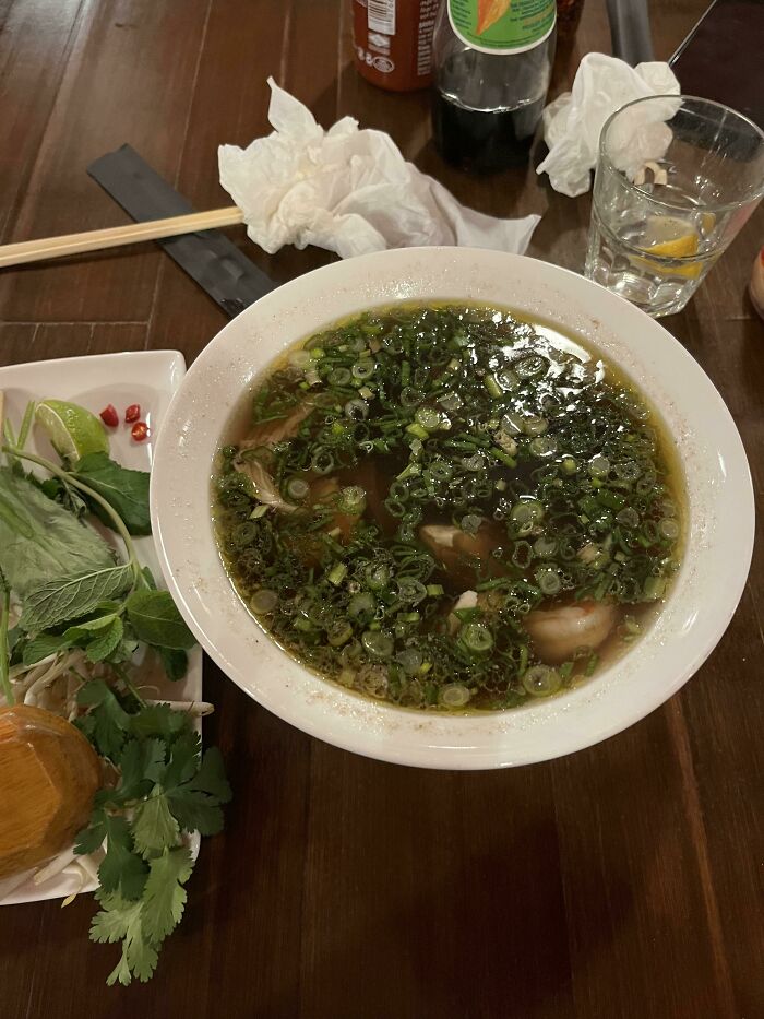 Sopa preparada con hierbas frescas al lado, junto a vaso con limón y condimentos sobre mesa de madera.