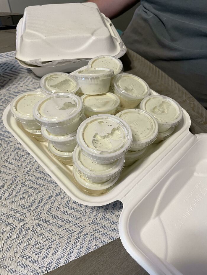 Vasos pequeños de salsa ranch en cantidades grandes dentro de caja de comida para llevar, personas alegres al recibir pedido exacto.