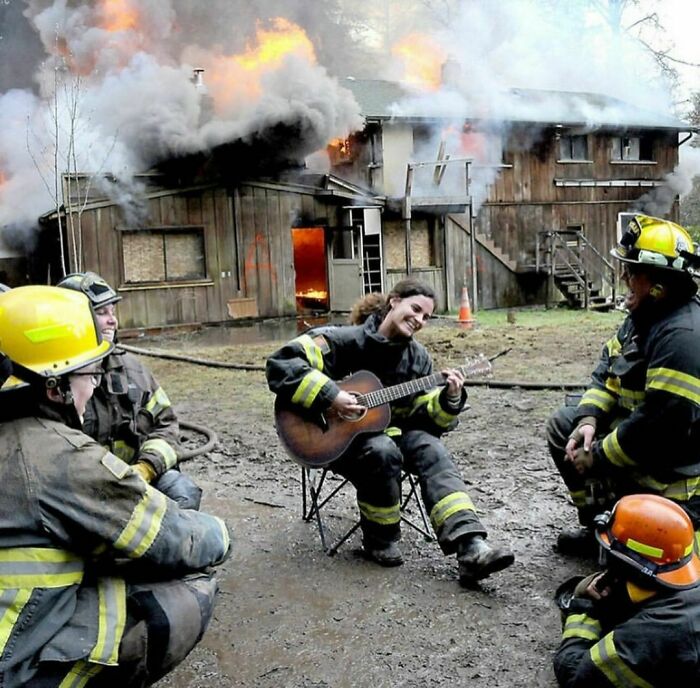 Bomberos sentados alrededor de una mujer tocando guitarra frente a una casa en llamas, imagen divertida y un poco incómoda.