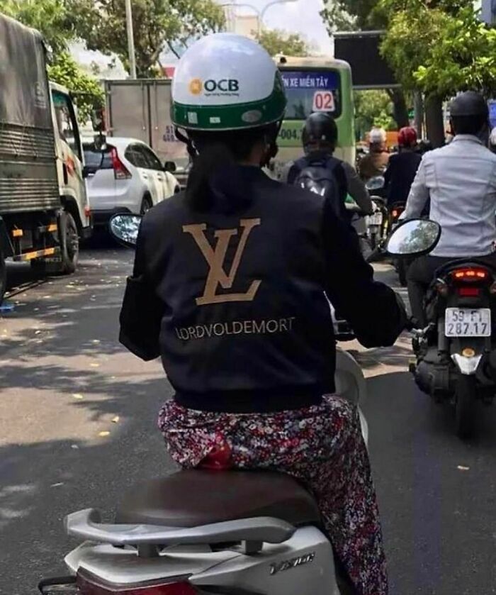 Persona en moto con casco y chaqueta negra con logo falso y texto divertido, en tráfico urbano, imagen incómoda y divertida.