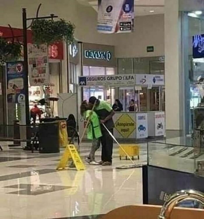 Pareja de empleados de limpieza abrazándose mientras trabajan en un centro comercial, imagen realmente incómoda pero divertida.