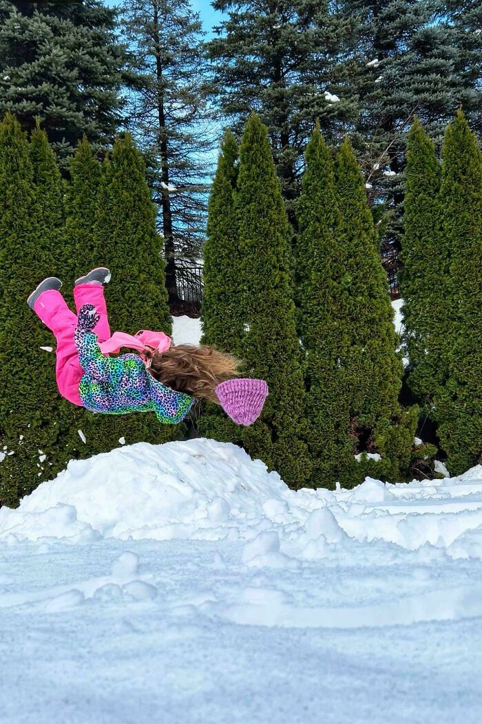 Niña haciendo una acrobacia en la nieve rodeada de árboles, refleja momentos divertidos y mejores de padres en familia.