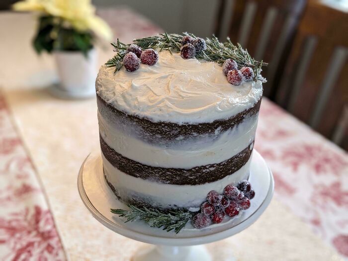 Pastel de repostería decorado con crema, arándanos y ramas verdes en base blanca sobre mesa con mantel.