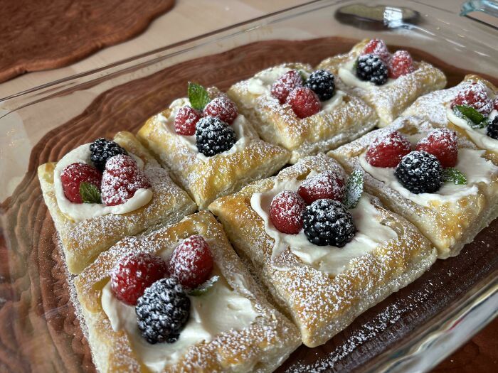 Repostería con hojaldre, crema y frutos rojos decorados con azúcar glas en un recipiente de vidrio sobre mantel marrón.