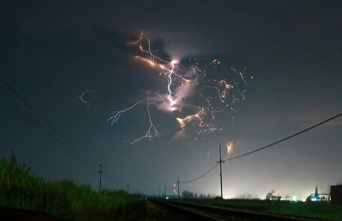 Rayo con efectos surrealistas ilumina el cielo nocturno en un paisaje rural con vías y pasto verde.