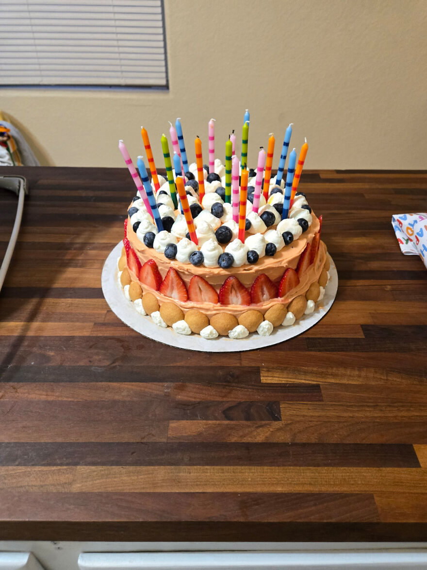 Pastel casero decorado con fresas, arándanos, crema y velas de colores sobre una mesa de madera oscura.