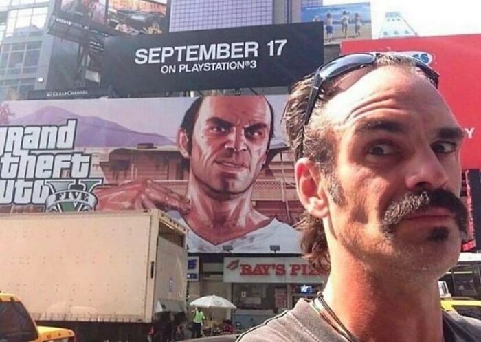 Hombre con bigote posando frente a cartel de Grand Theft Auto V en una calle concurrida, imagen divertida viral.