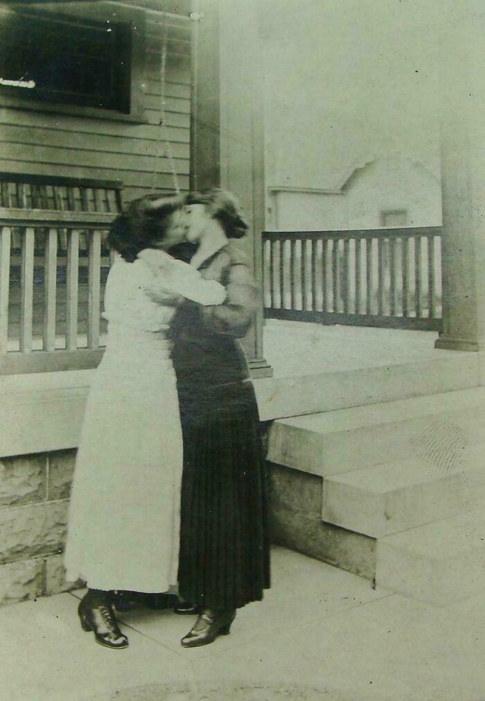 Dos mujeres victorianas besándose frente a una casa antigua con vestimentas típicas de la época victoriana.