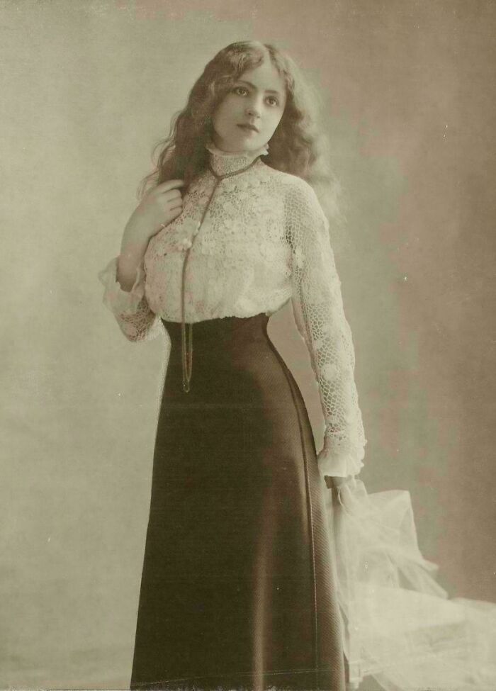 Mujer victoriana con blusa de encaje y falda larga sosteniendo velo, mostrando moda de la época victoriana.