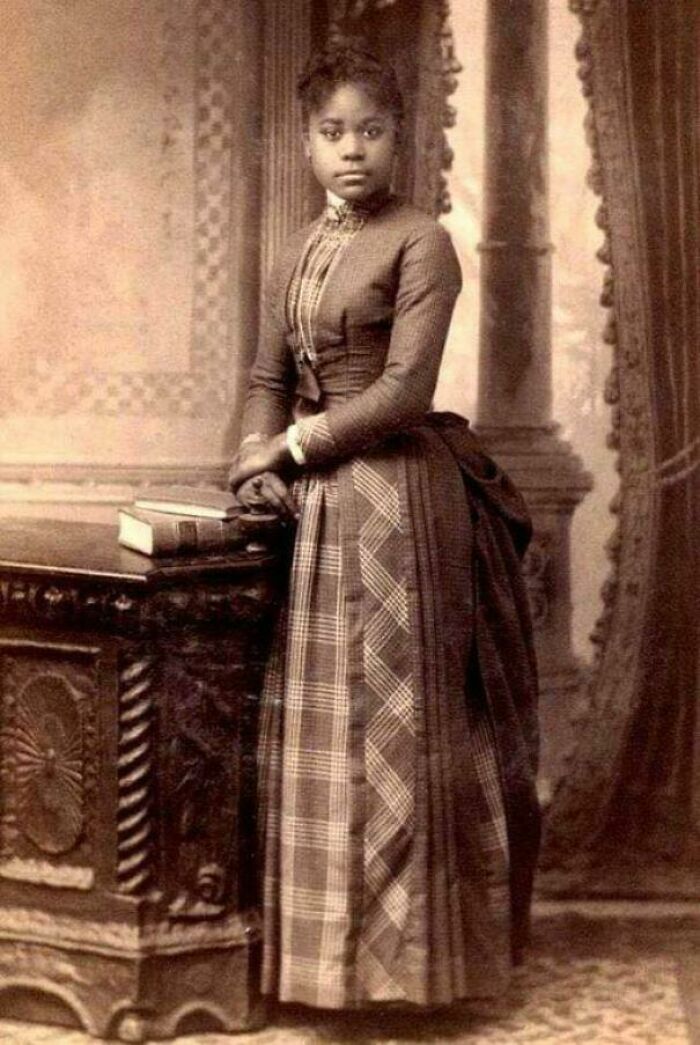 Mujer posando en época victoriana con vestido largo y libro sobre mesa tallada mostrando tiempos muy distintos.
