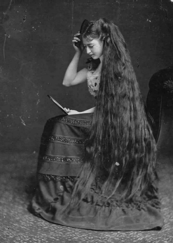 Joven victorian en vestido largo con cabello muy extenso, sosteniendo un sombrero y leyendo un libro en blanco y negro.