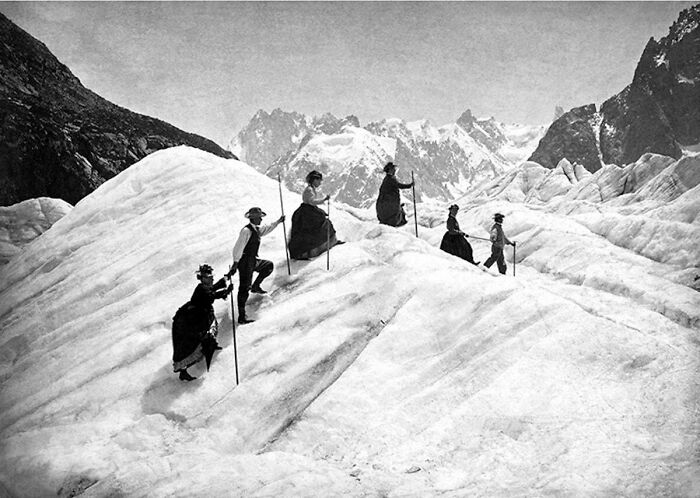 Personas con ropa de la época victoriana caminando sobre un glaciar en un paisaje montañoso nevado.