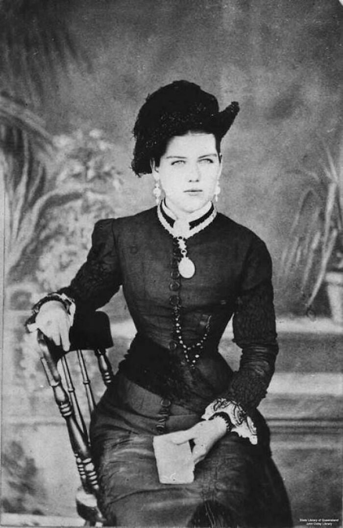 Mujer victoriana sentada con vestido oscuro elegante y sombrero, mostrando moda típica de la época victoriana en blanco y negro.