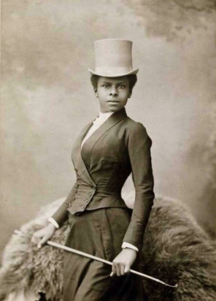 Mujer vestida con traje victoriano y sombrero alto, posando con bastón en imagen histórica de la época victoriana.
