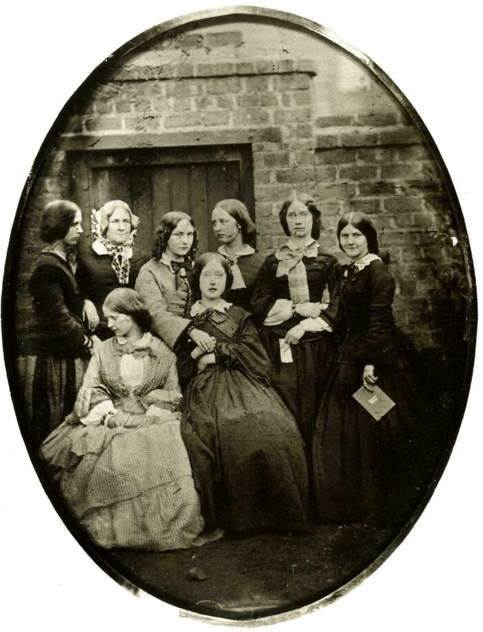 Grupo de mujeres en época victoriana posando frente a una pared de ladrillos, mostrando vestimenta típica victoriana.