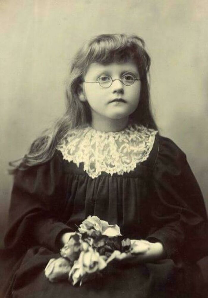 Niña con gafas y vestido antiguo sosteniendo flores en una fotografía histórica de la época victoriana.