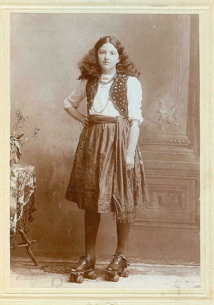 Joven con ropa de la época victoriana posando con patines de cuatro ruedas en estudio fotográfico antiguo.