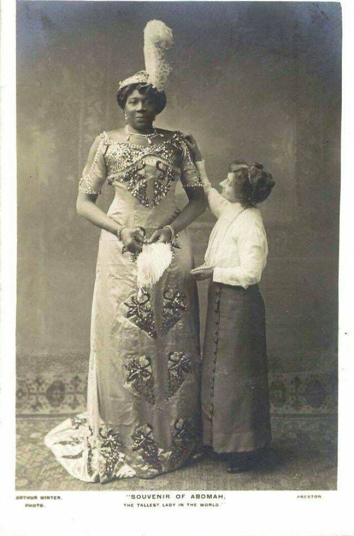 Mujer alta con vestido victoriano adornado y otra persona ajustando su atuendo en imagen histórica época victoriana.