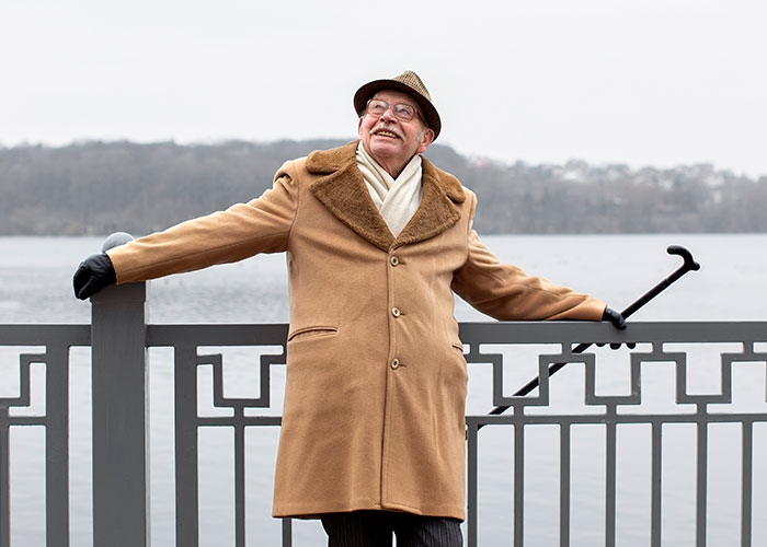 Hombre mayor con abrigo marrón y bastón, sonriendo junto a un lago, evocando secretos familiares ocultos.