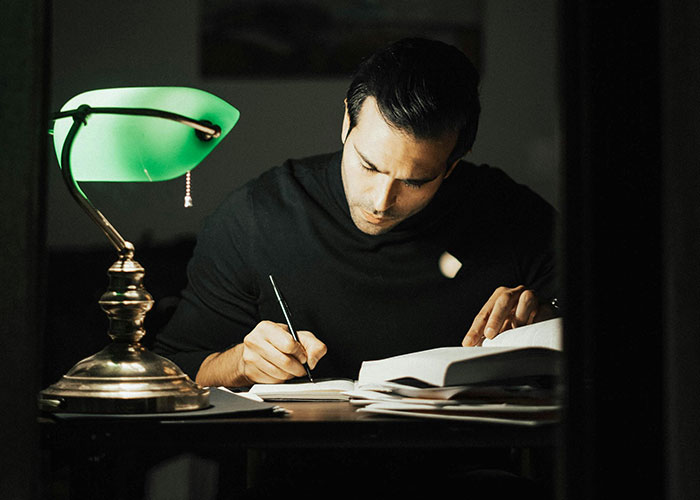 Hombre escribiendo bajo luz de lámpara verde, evocando secretos familiares misteriosos.