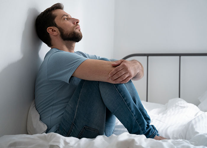 Hombre pensativo en la cama, vestido con camiseta y jeans, evocando secretos familiares.