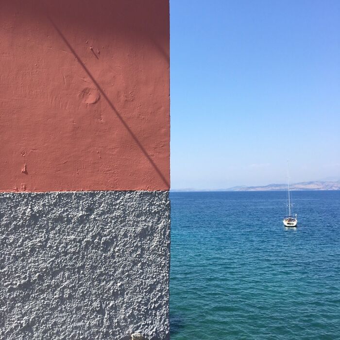 Mitad de pared dividida en colores contrastantes frente a un mar con barco, imagen con toque de surrealismo sorprendente.
