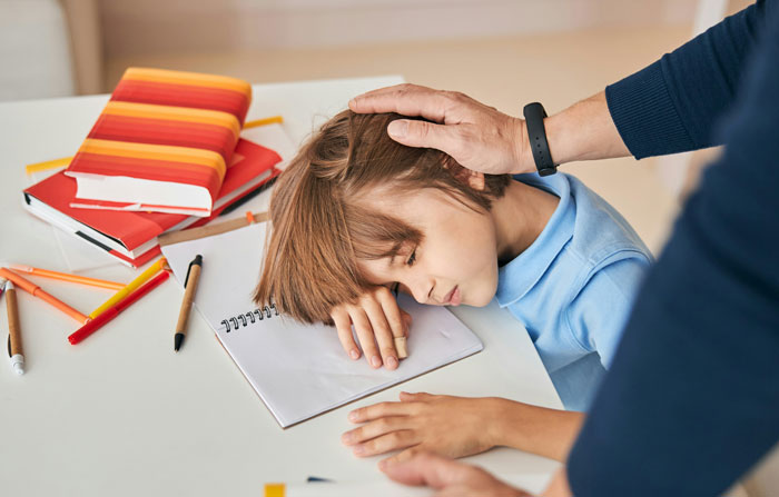 Alumno durmiendo sobre un cuaderno en clase, rodeado de libros y bolígrafos, mientras un profesor lo acaricia.
