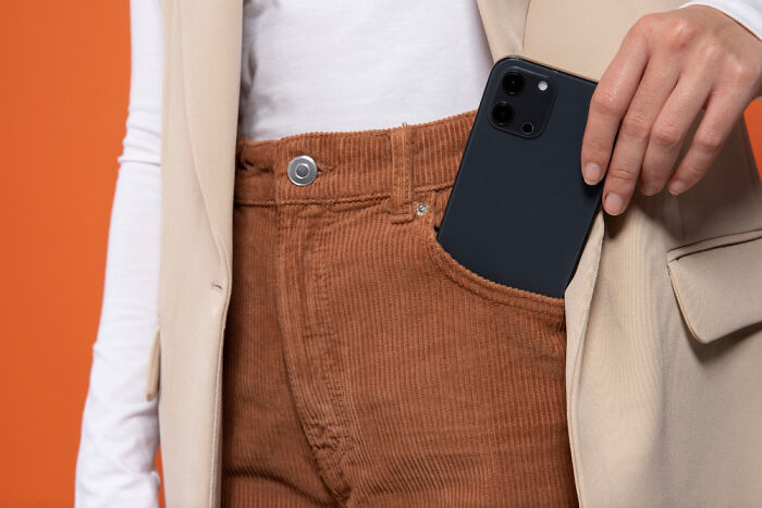 Persona guardando un teléfono móvil en el bolsillo de un pantalón de pana marrón con chaqueta beige sobre fondo naranja.