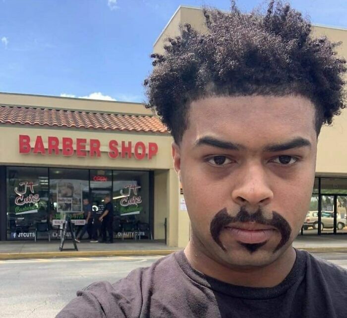 Hombre con corte de cabello y bigote peculiar frente a barbería, imagen divertida que causa risas entre millones de personas.