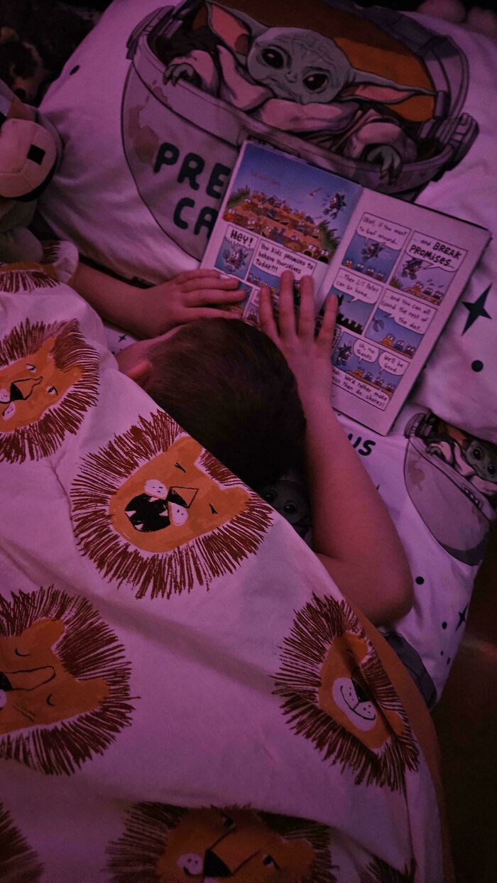 Niño acostado leyendo un libro en cama con prendas de leones y personajes, momento entrañable de paternidad.