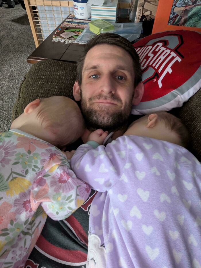 Padre sonriente recostado con dos bebés durmiendo sobre su pecho en momento entrañable y divertido de paternidad.