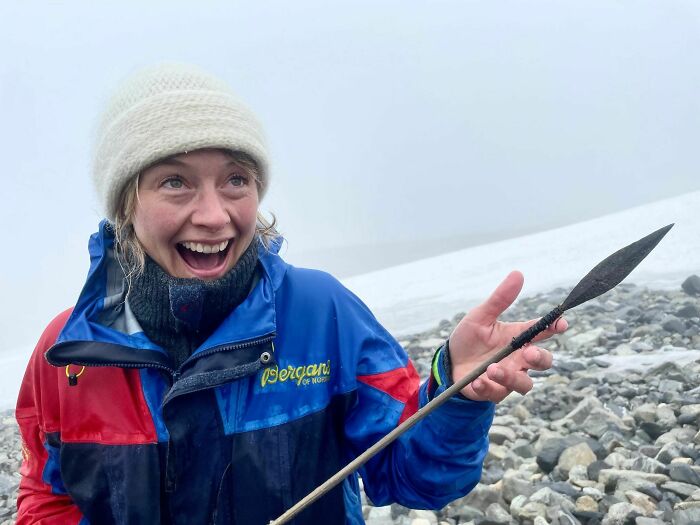 Mujer sonriente en prenda de invierno sostiene punta de lanza antigua, hallazgo fascinante del pasado lejano.