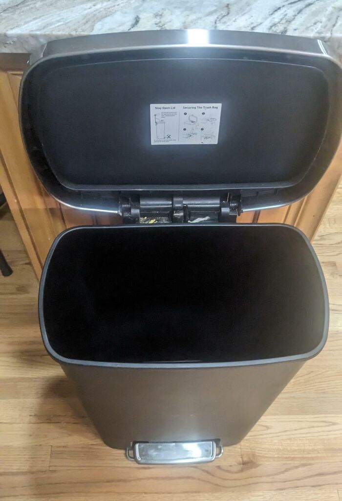 Un bote de basura vacío con tapa abierta en una cocina, destacando ejemplos de novios y maridos.