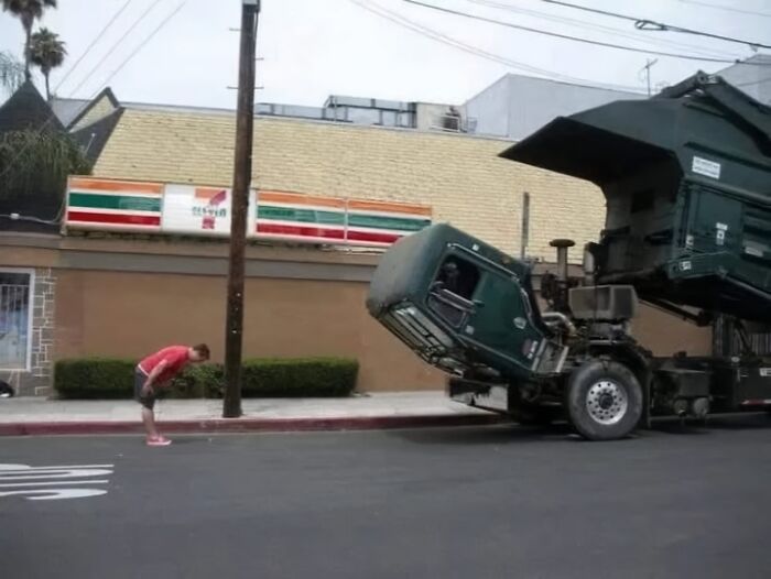 Persona observando un camión de basura levantado en una posición inusual frente a una tienda.