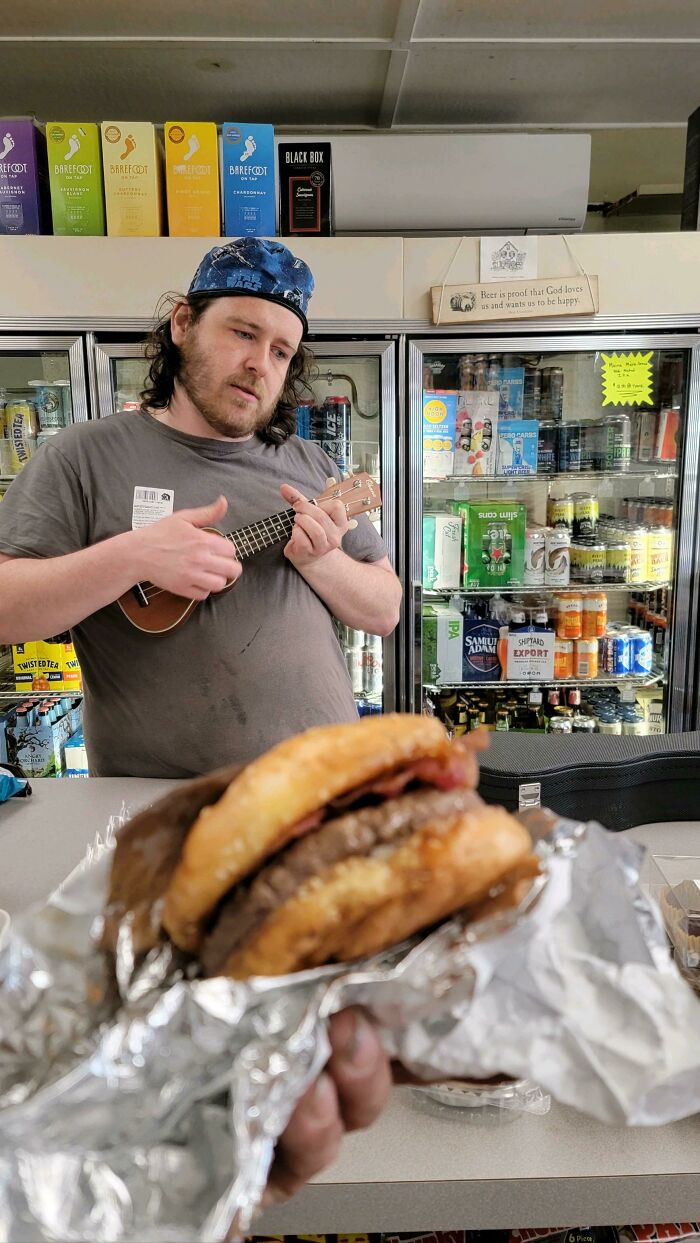 Empleado de hostelería tocando un ukelele frente a un refrigerador, con una hamburguesa en primer plano.