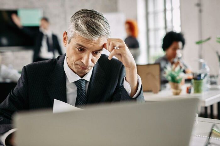 Hombre de negocios preocupado leyendo documentos en oficina moderna, reflejando estupideces y confusión laboral.