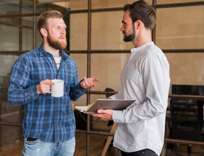 Dos hombres conversando en oficina informal, uno con taza de café y otro con portapapeles, mostrando comunicación casual y expresiva.