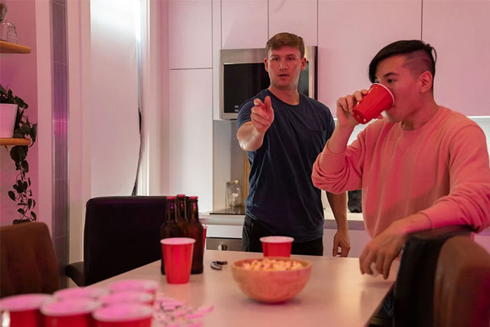 Dos personas en una fiesta en casa, una señalando y otra bebiendo de un vaso rojo, reflejando la necesidad de irse.