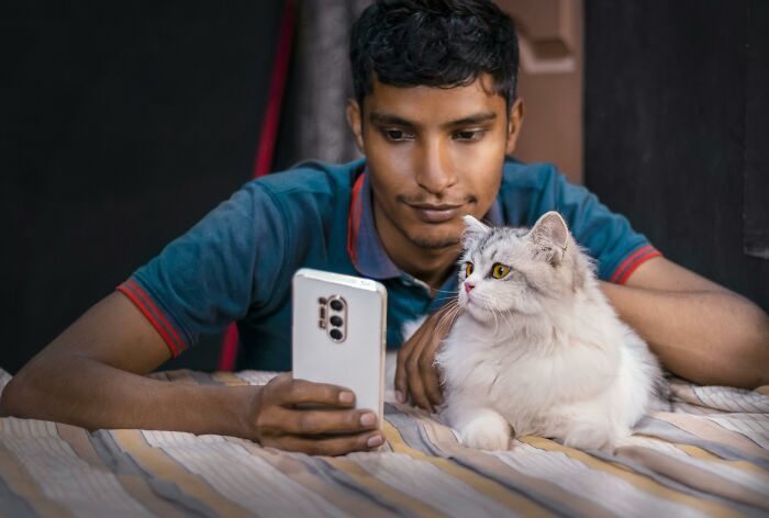Joven con camisa azul mirando un teléfono junto a un gato esponjoso, posiblemente escuchando conversaciones accidentales.