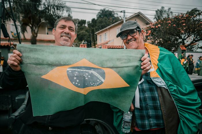 Dos hombres sonrientes sostenien una bandera brasileña en una reunión al aire libre.