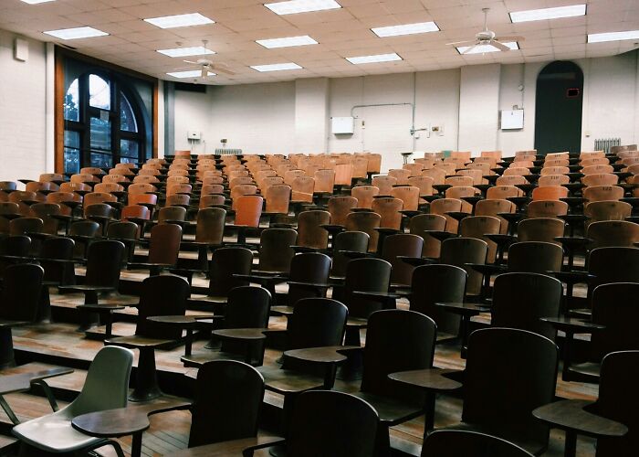 Aula vacía con asientos de madera, escenario de conversaciones escuchadas accidentalmente.