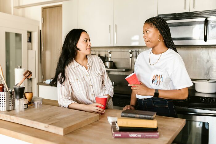 Dos mujeres conversando en una cocina, cada una con un vaso rojo, en un ambiente casual.