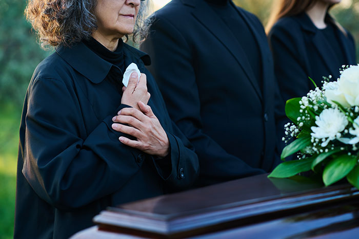 Personas de luto junto a un ataúd y flores blancas, en un funeral al aire libre.