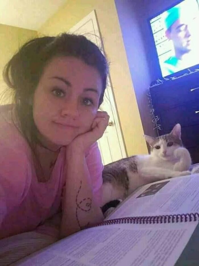 Persona descansando junto a un gato, ambos mirando a cámara y un televisor al fondo.