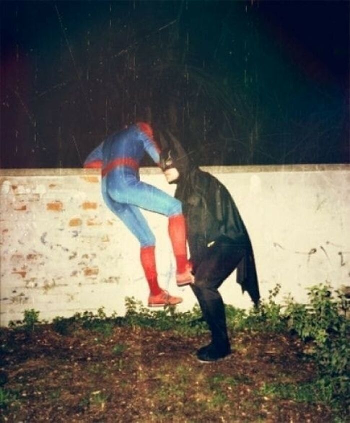 Dos personas disfrazadas de superhéroes, realizando una acción inusual junto a un muro, en una escena rara y absurda.