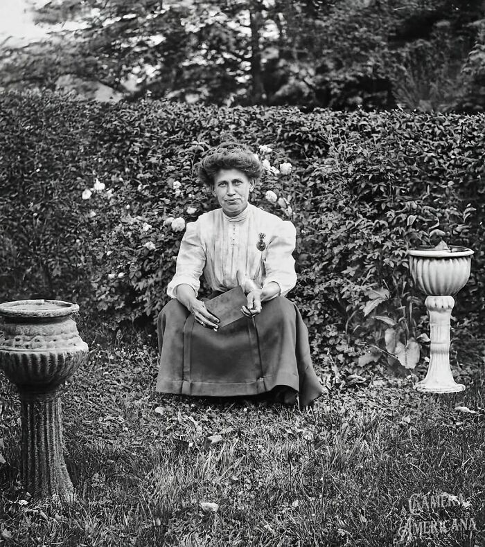 Mujer sentada en un jardín, rodeada de vegetación, sosteniendo un libro, evocando tiempos históricos pasados.